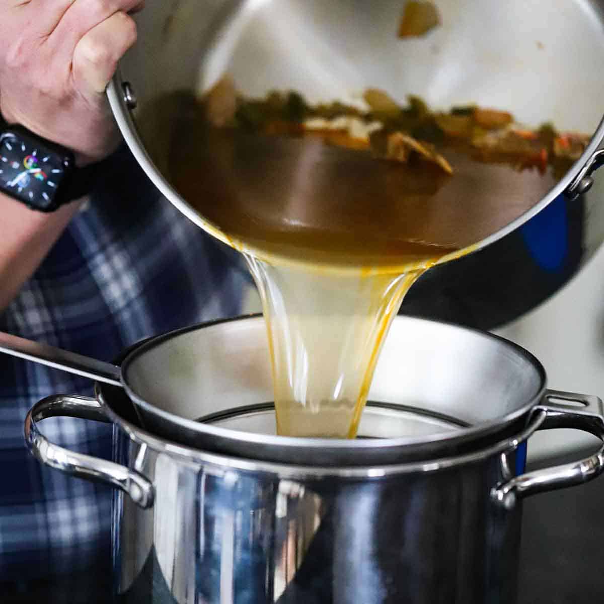 A person straining homemade shellfish stock from a large stock pot into another pot lined with a fine-mesh sieve.