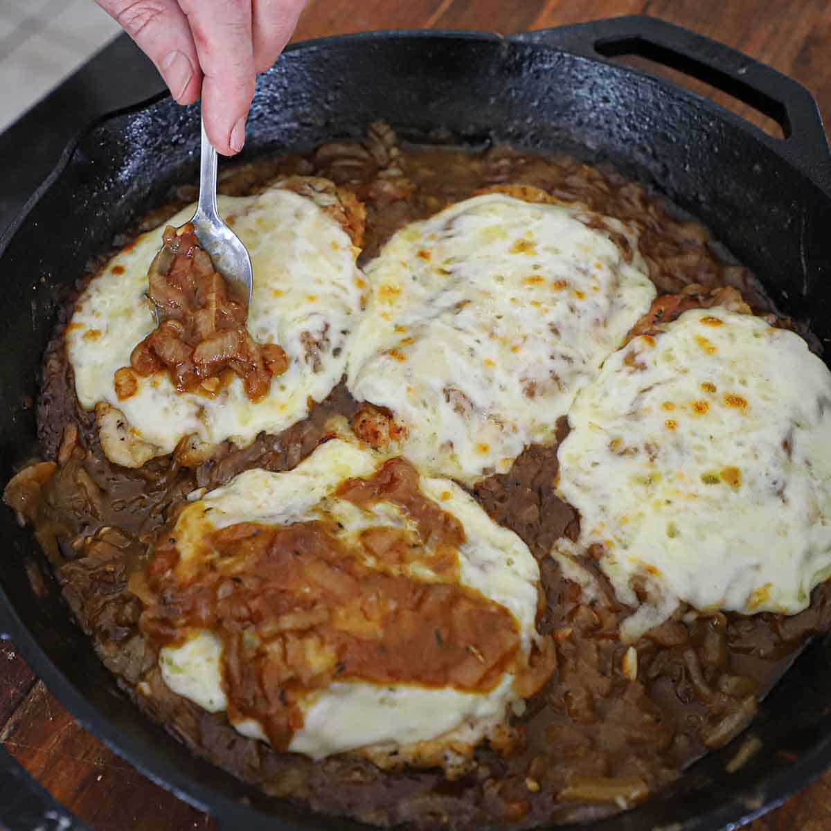 A person spoon French onion sauce over the top of a French onion smothered chicken that is resting in a pan with three other smothered chicken breasts all in a cast-iron skillet.