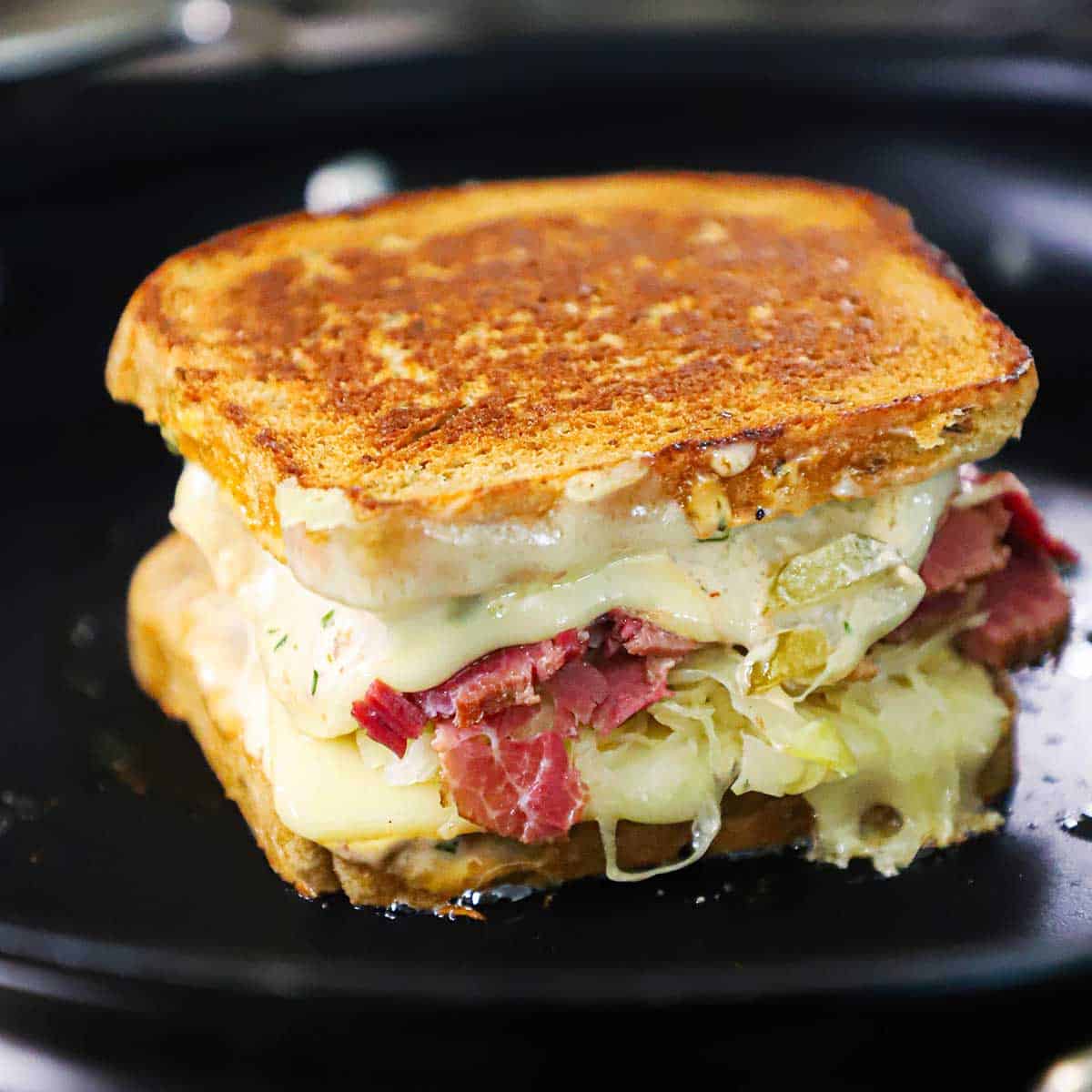 A "from scratch" reuben sandwich that has melty cheese and piles of corned beef and sauerkraut being grilled in a non-stick skillet.