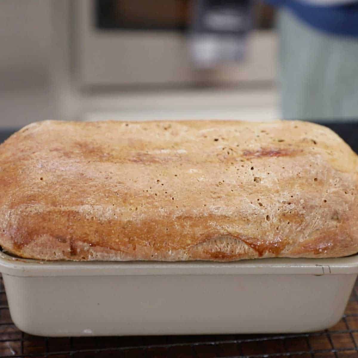 A freshly baked deli-style rye bread loaf that is still in the loaf pan and is resting on a wire rack on a wooden cutting board.