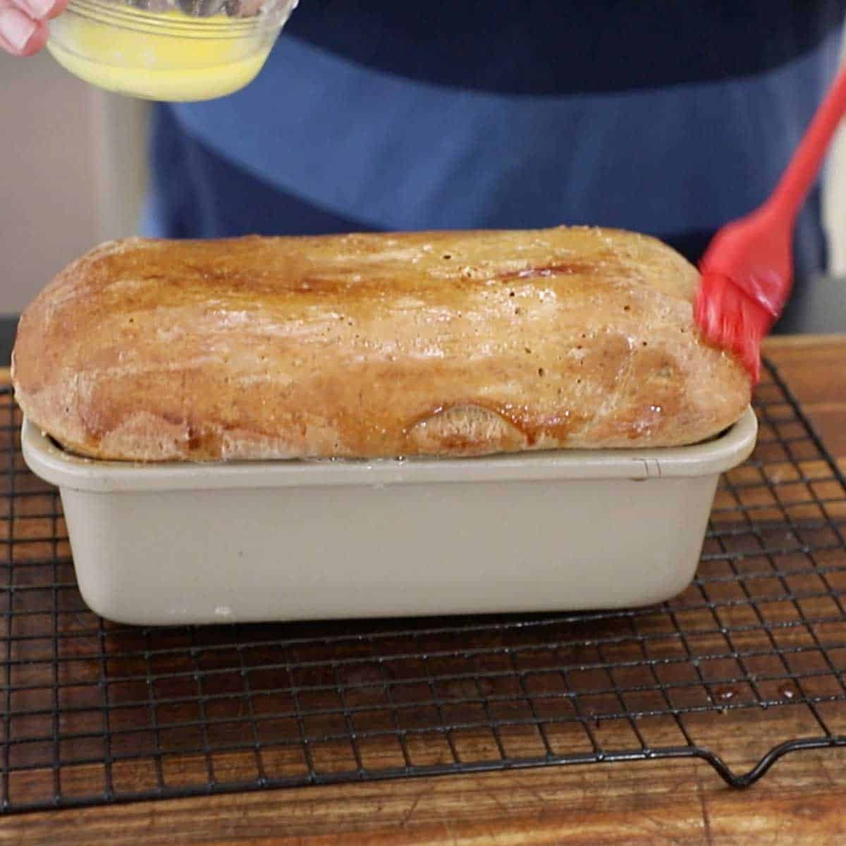 A person brushing melted butter all over the top of a deli-style rye bread loaf that is in a loaf pan on a baking rack on a wooden cutting board.