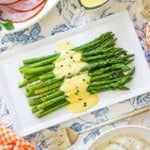 An overhead view of a white rectangle platter filled with a dish of Asparagus with Béarnaise sauce and a large gold serving fork nearby.