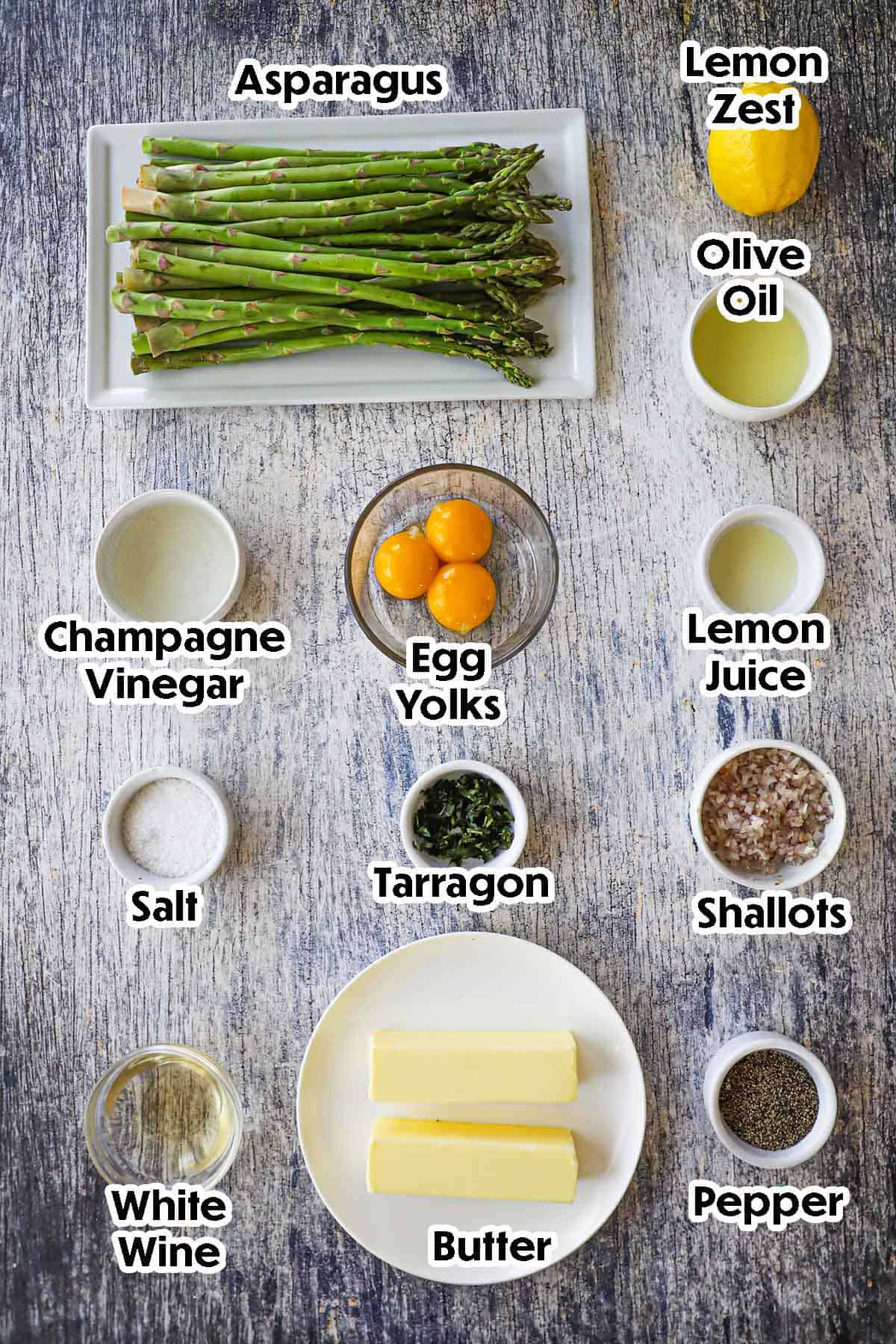 An arrangement of ingredients for Asparagus with Béarnaise Sauce on a grey wooden background including asparagus, egg yolks, tarragon, vinegar, lemon juice, shallots, butter, wine, and seasonings.