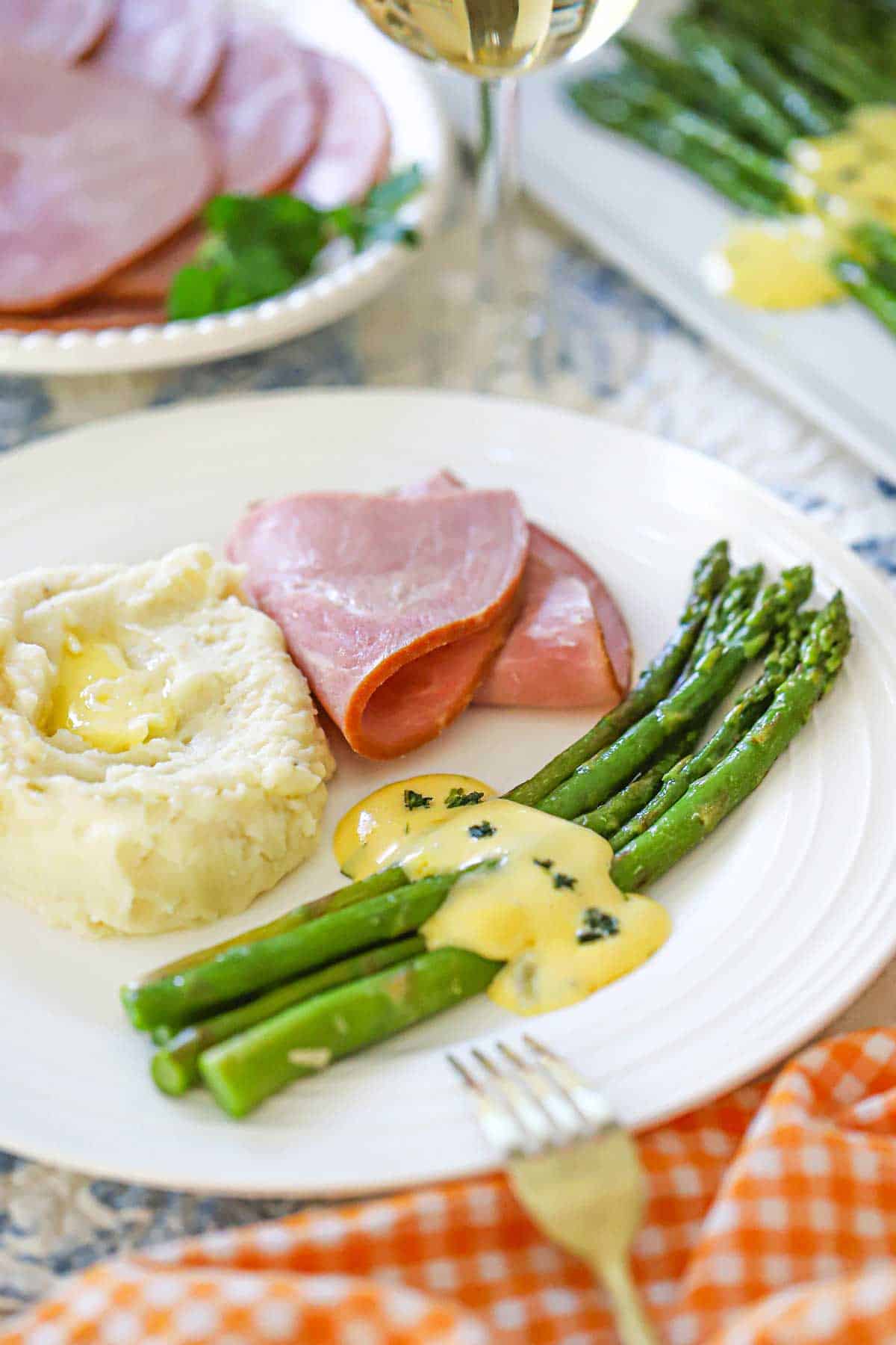 A white dinner plate filled with a single serving of asparagus with Béarnaise sauce next to a pile of mashed potatoes and a couple slices of ham.