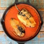 An overhead view of two chile rellenos resting in a red sauce in a large cast-iron skillet.