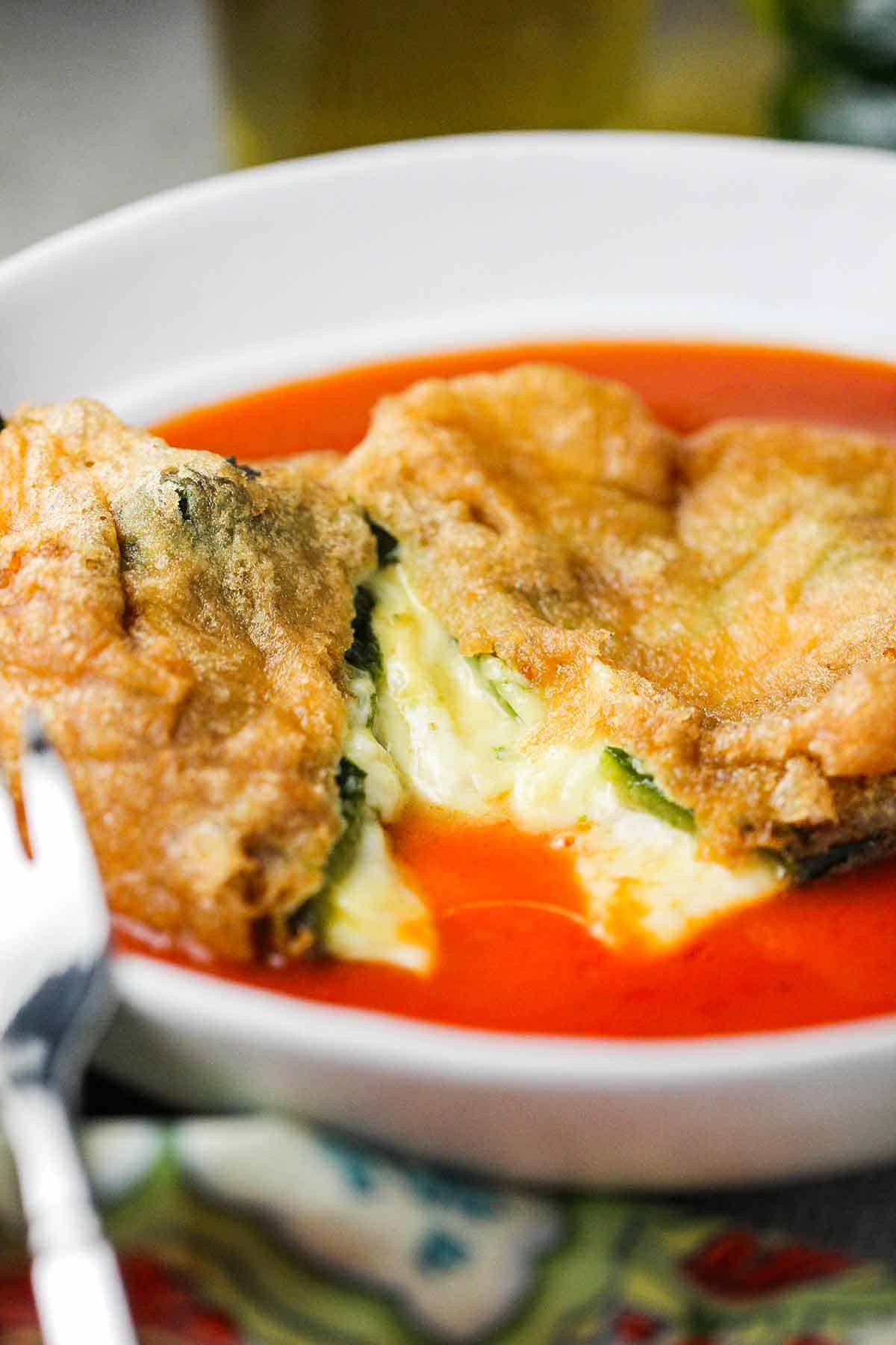 A straight-on view of an authentic chile relleno that has been split open with melted cheese oozing out into the bowl that contains a thin layer of red sauce.