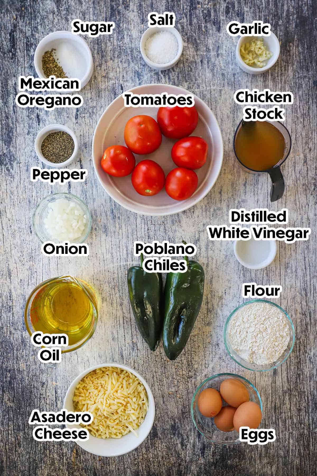 An arrangement of ingredients for Authentic Chile Relleno on a wooden grey background including poblano chiles, cheese, flour, eggs, onion, vinegar, oil, chicken stock, garlic, tomatoes, and seasonings.