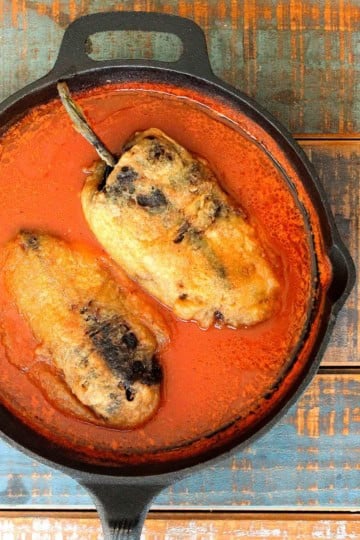 An overhead view of two chile rellenos resting in a red sauce in a large cast-iron skillet.