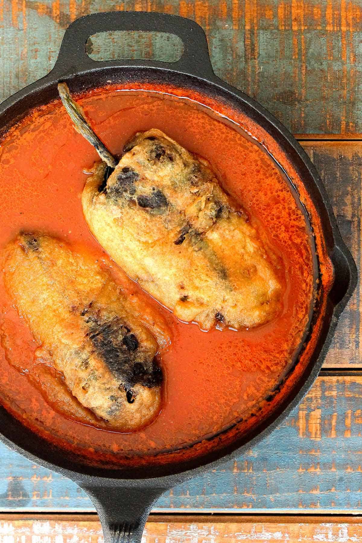 An overhead view of two chile rellenos resting in a red sauce in a large cast-iron skillet.