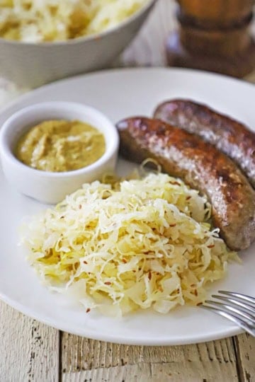 A close-up view of a pile of homemade sauerkraut on a white dinner plate with two seared bratwurst links and a small bowl of brown mustard sitting on the plate nearby.