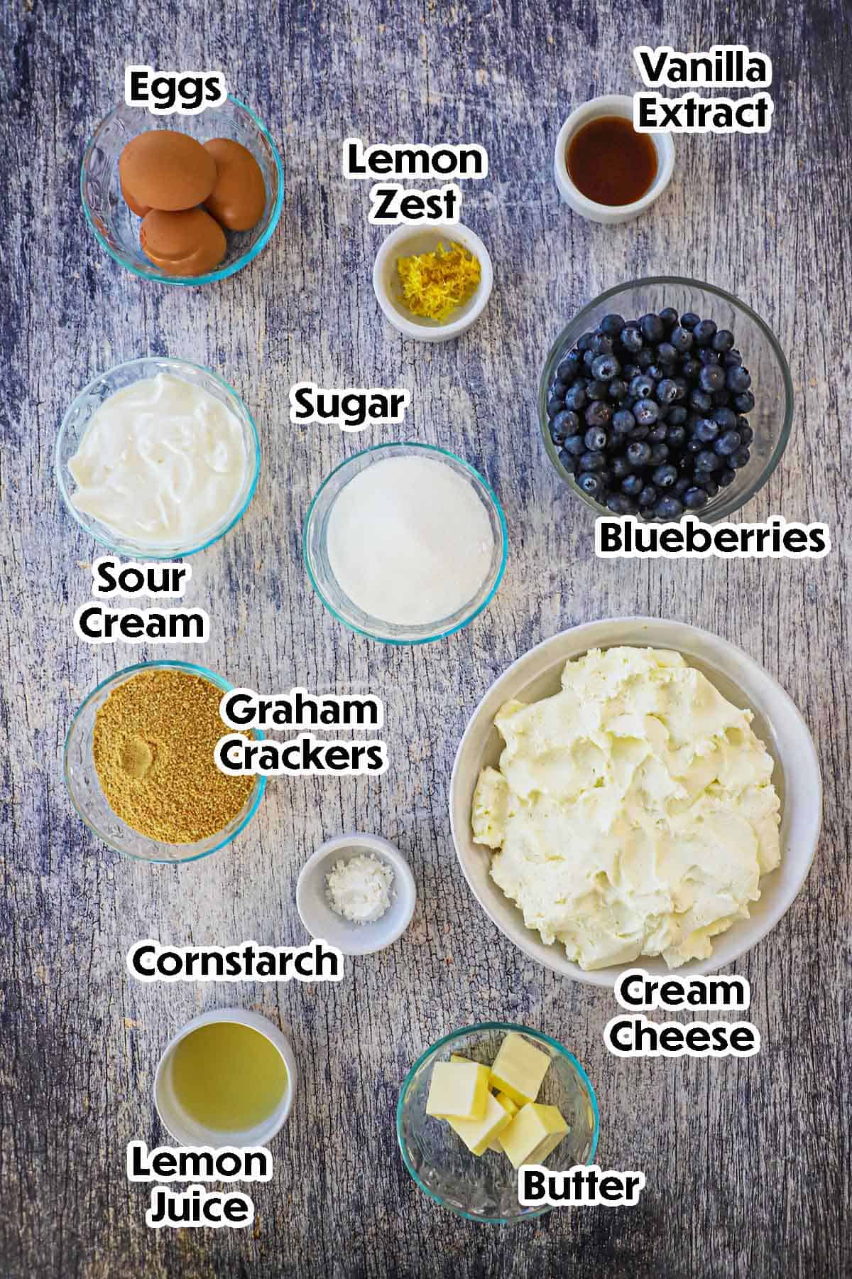 An arrangement of ingredients for lemon blueberry cheesecake on a grey wooden background including softened cream cheese, blueberries, eggs, sugar, lemon zest, vanilla, graham crackers, butter, and lemon juice.