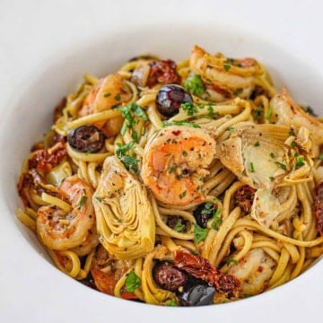 A close-up view of a large white pasta bowl filled with Mediterranean past with shrimp garnished with chopped Italian parsley.