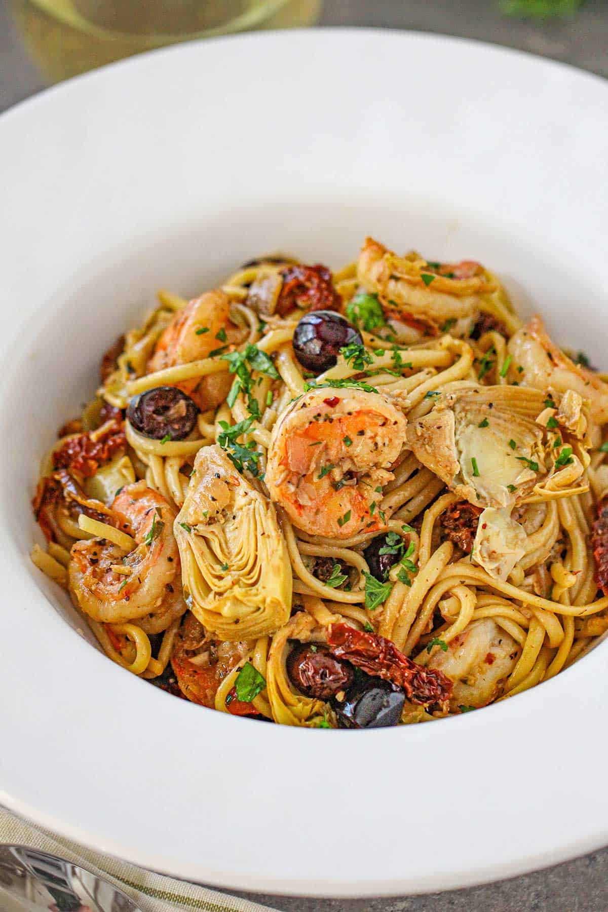 A close-up view of a large white pasta bowl filled with Mediterranean past with shrimp garnished with chopped Italian parsley.