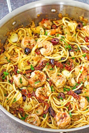 An overhead view of a large stainless steel skillet filled with Mediterranean pasta with shrimp topped with chopped Italian parsley.