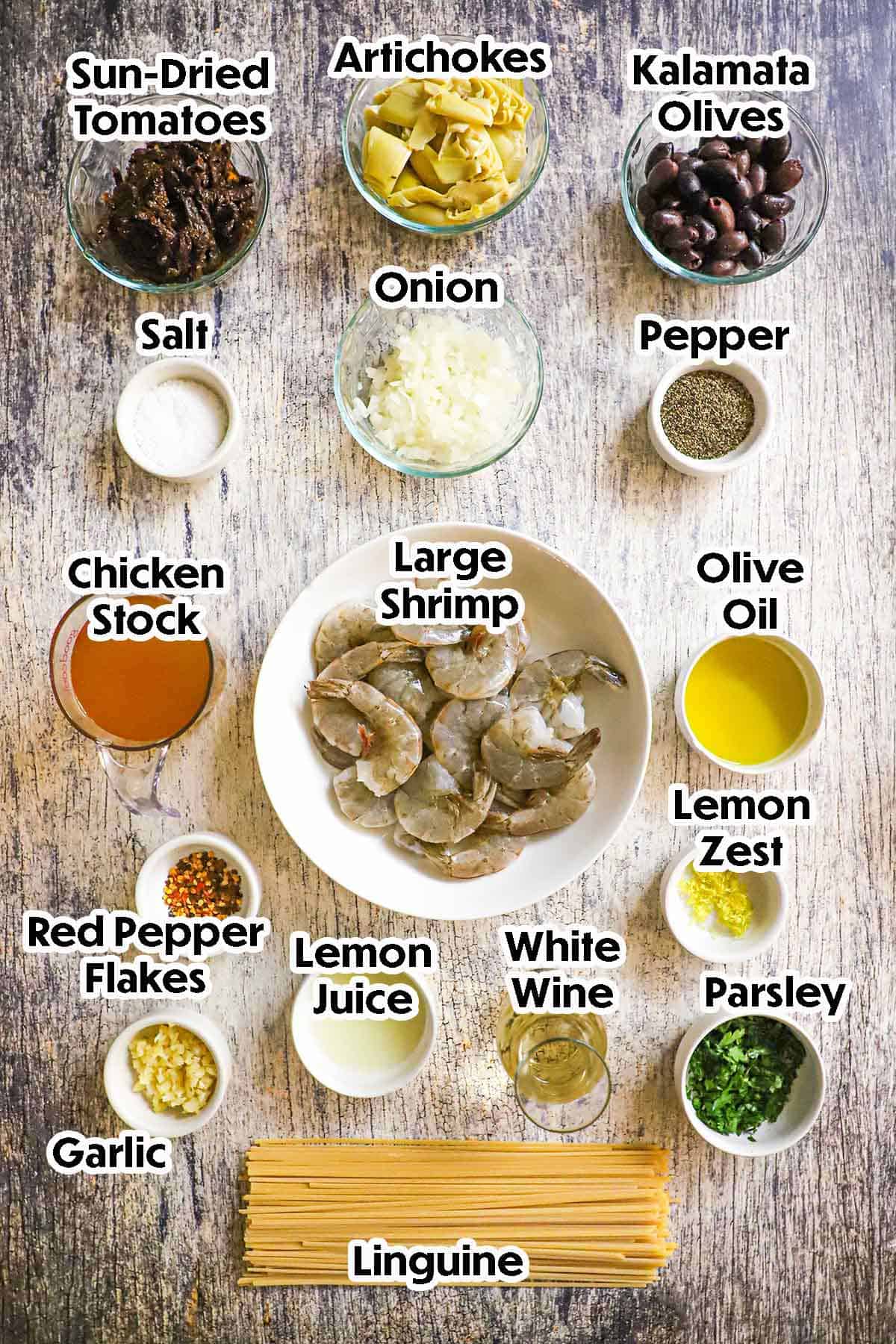 An arrangement of ingredients for Mediterranean Pasta with Shrimp on a grey wooden background including raw shrimp, sun-dried tomatoes, artichokes, olives, onion, chicken stock, oil, garlic, lemon juice, white, wine, pasta, herbs, and seasonings.