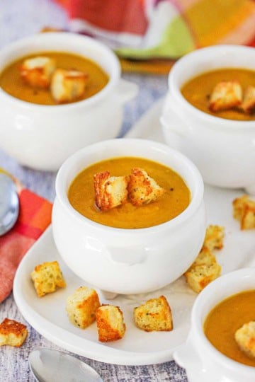 Four white soup crocks filled with roasted butternut squash soup topped with homemade croutons with more croutons scattered around the base of the bowls.