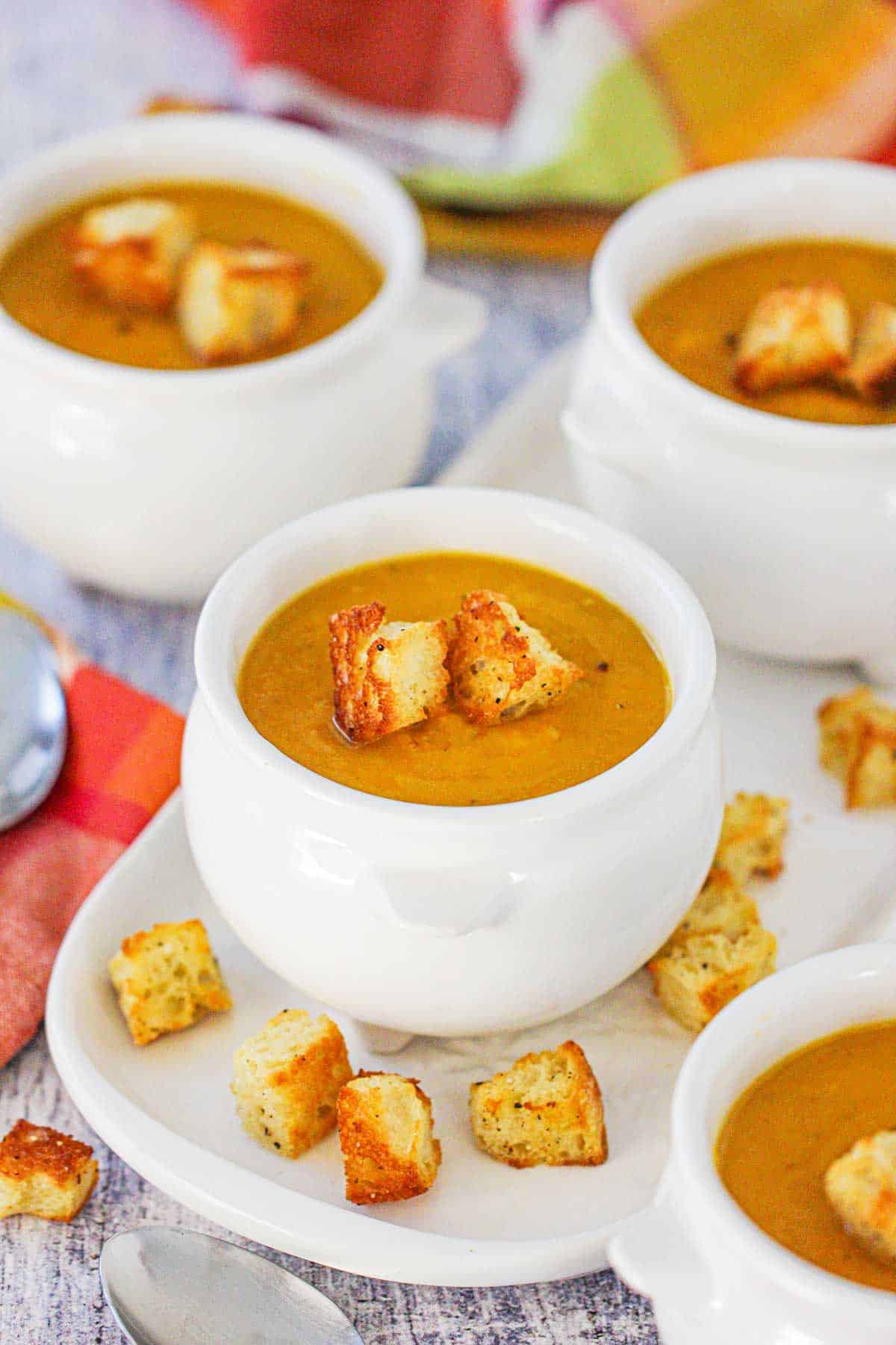 Four white soup crocks filled with roasted butternut squash soup topped with homemade croutons with more croutons scattered around the base of the bowls.