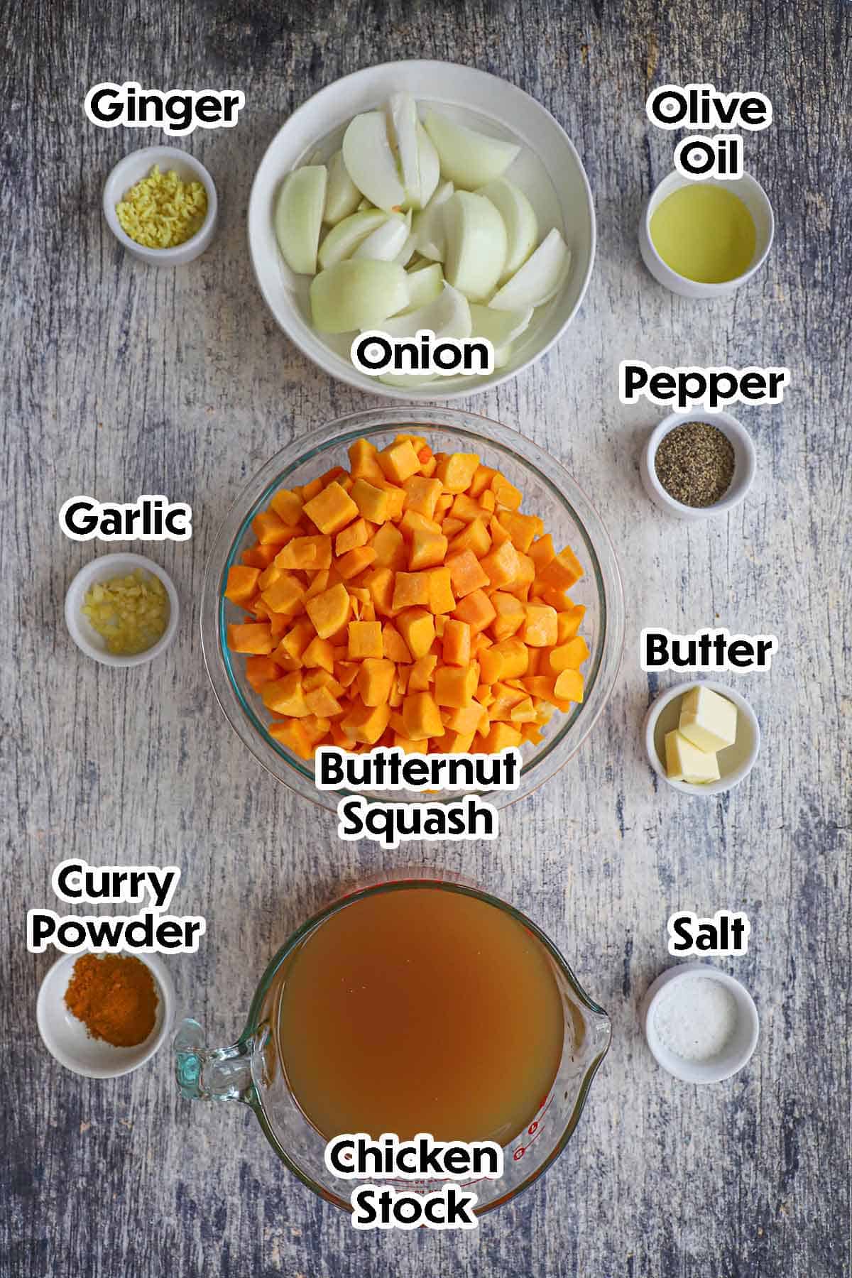 An arrangement of ingredients for roasted butternut squash soup on a grey wooden background including cubed butternut squash, onion, ginger, chicken stock, garlic, butter, oil, and seasonings.