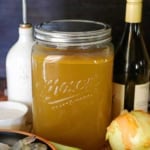 A close-up view of an extra-large Mason glass jar filled with homemade shellfish stock and is surrounded by a bowl filled with uncooked shrimp, leeks, onion, and a bottle of white wine.
