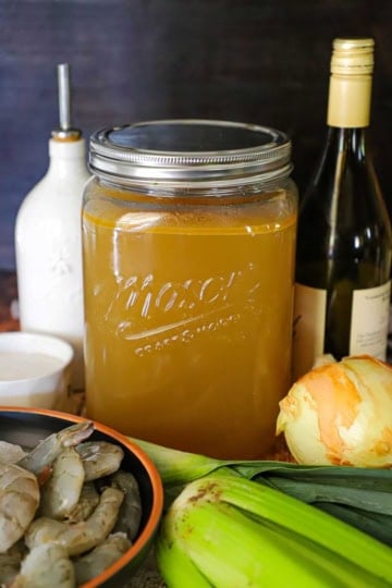 An extra-large Mason glass jar filled with homemade shellfish stock and is surrounded by a bowl filled with uncooked shrimp, leeks, onion, and a bottle of white wine.