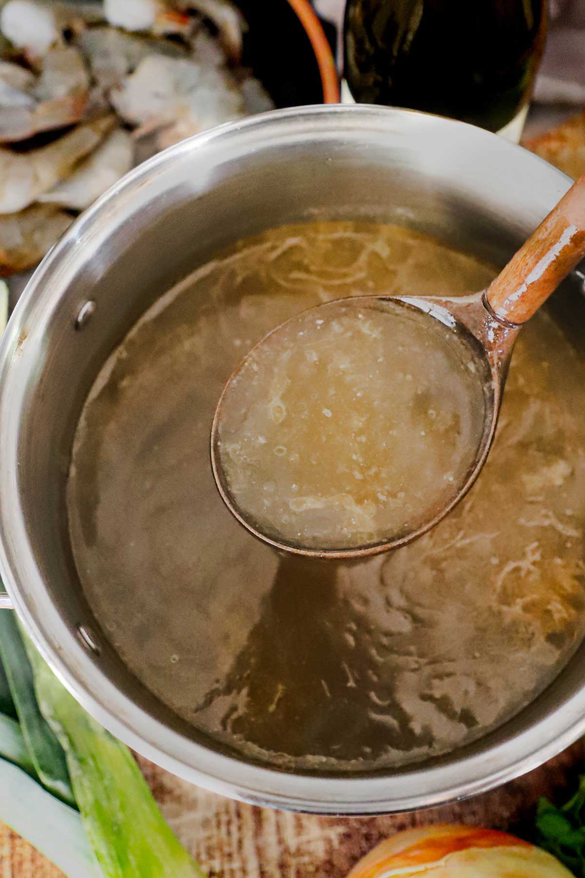 A person using a large wooden ladle to raise a ladleful of homemade shellfish stock from a stock pot filled with the stock.