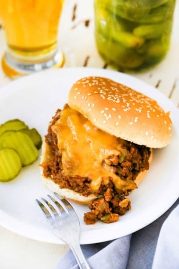 A homemade sloppy Joe sitting on a white dinner plate with a small pile of dill pickle chips next to the sandwich.