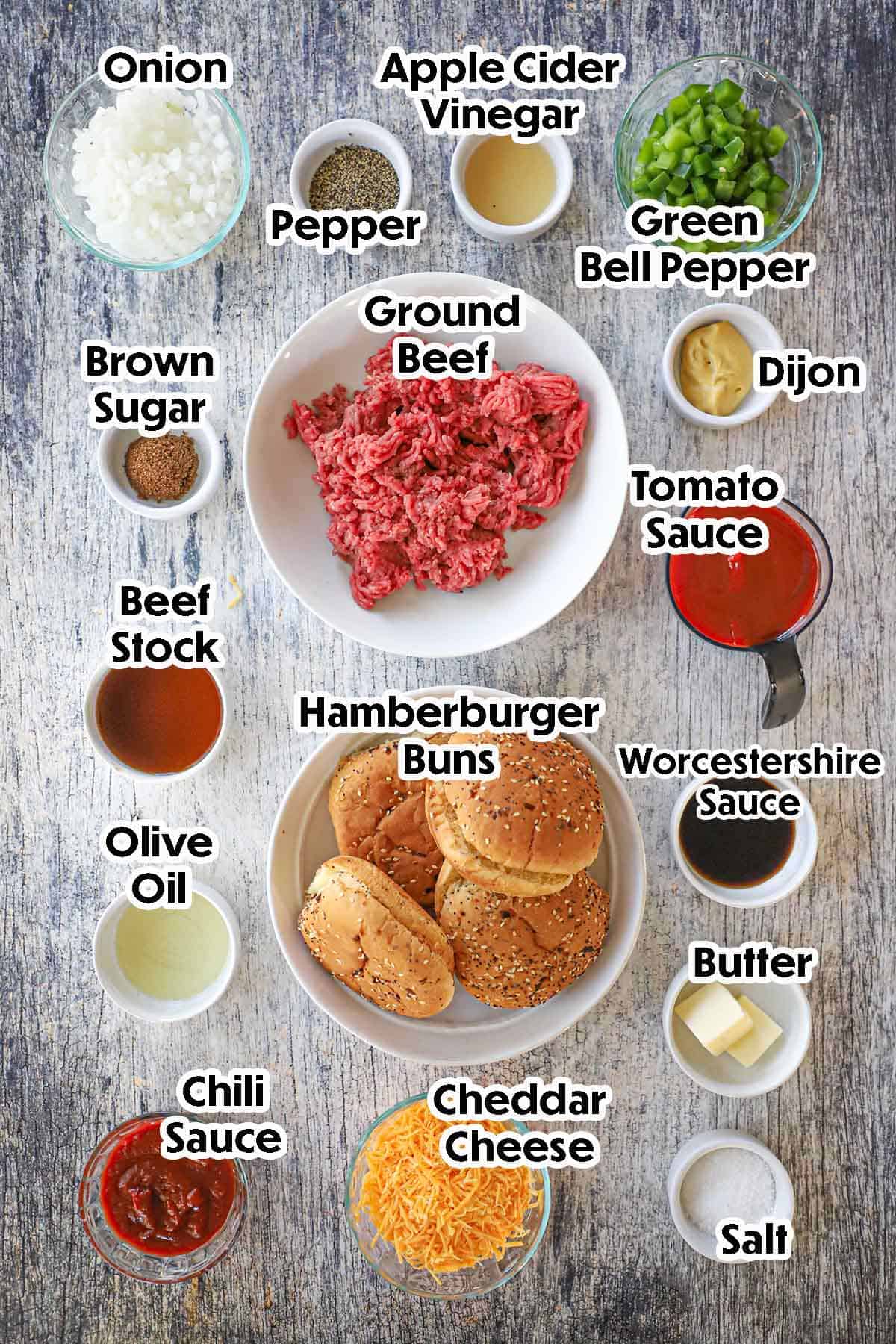 An arrangement of ingredients for homemade sloppy Joes on a grey wooden background including ground beef, onion, bell pepper, beef stock, tomato sauce, Worcestershire sace, chili sauce, hamburger buns, cheese, and seasonings.
