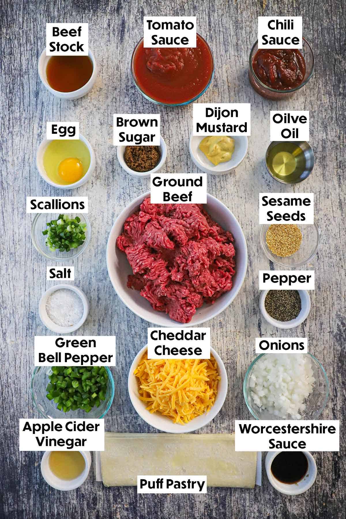 An arrangement of ingredients for Sloppy Joe Casserole on a grey wooden background including ground beef, tomato sauce, beef stock, chili sauce, egg, mustard, brown sugar, olive oil, cheese, onions, bell peppers, puff pastry, and seasonings.