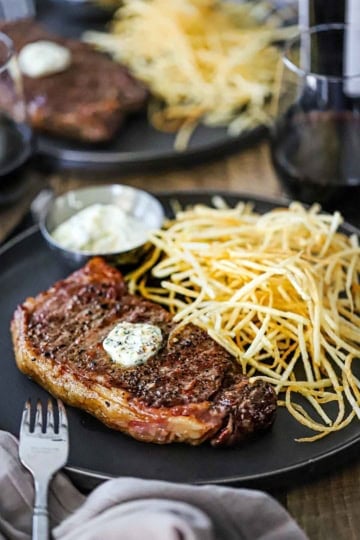 A straight-on view of French bistro-style Steak Frites on a large black plate including a reverse-seared strip steak and a pile of shoestring French fries next to a small bowl of garlic basil aioli.