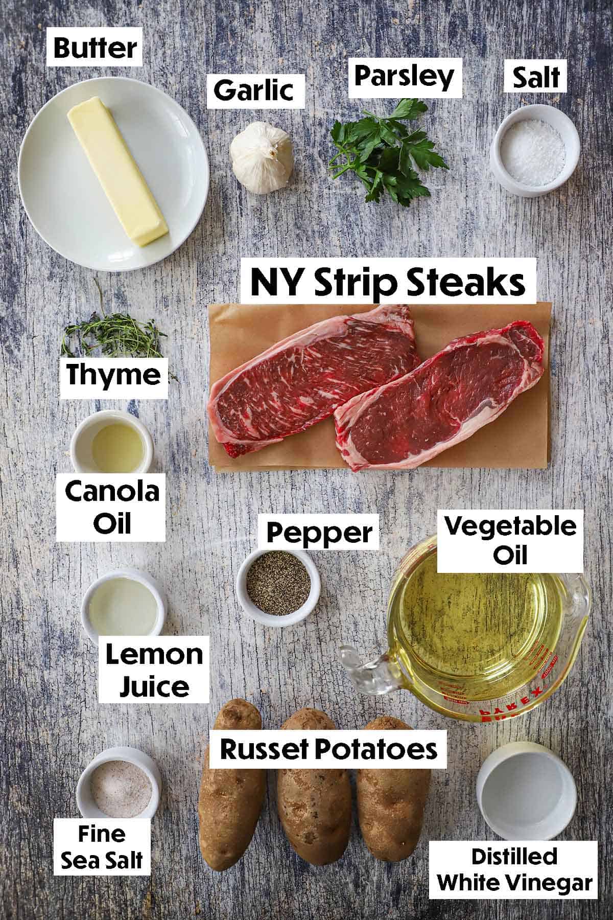 An arrangement of ingredients for steak frites on a grey wooden background including strip steaks, potatoes, butter, garlic, parsley, thyme, oil, lemon juice, vinegar, and seasonings.