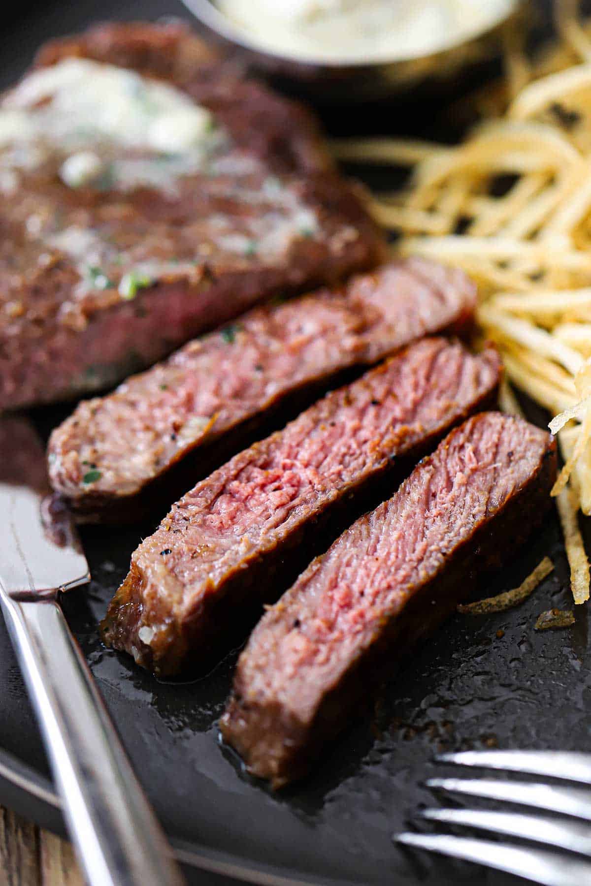 A close-up view of a reverse-seared New York strip steak that has been cut into strips sitting on a plate next to shoestring French fries.