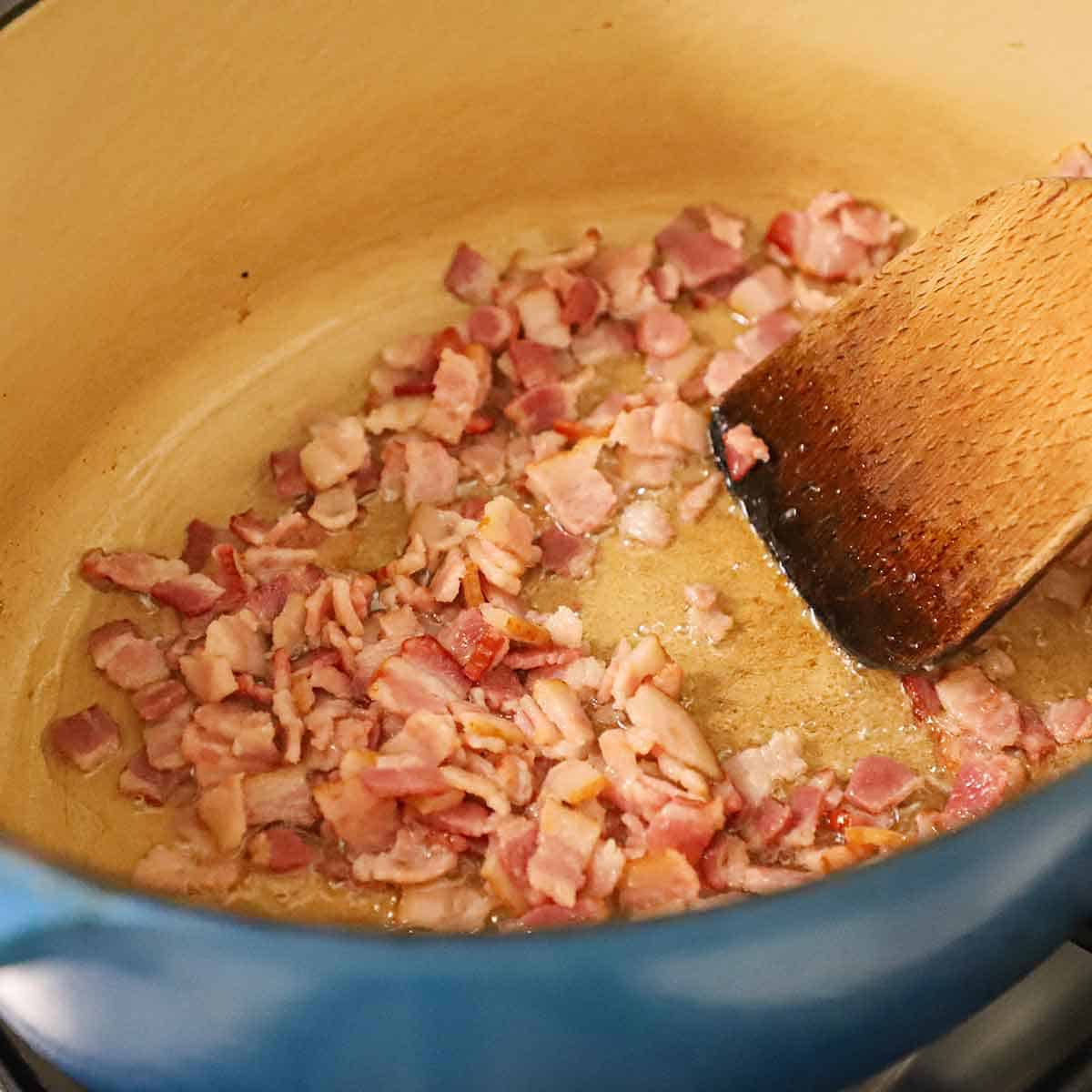A person using a wooden spatula to stir bacon pieces that are being cooked in large Dutch oven on a gas stove.