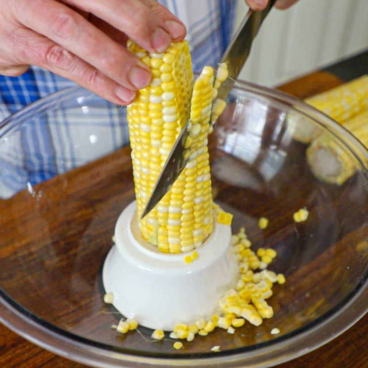 A person using a chef's knife to cut away kernels from a cob of corn that is resting on an inverted small bowl inside a large glass bowl.