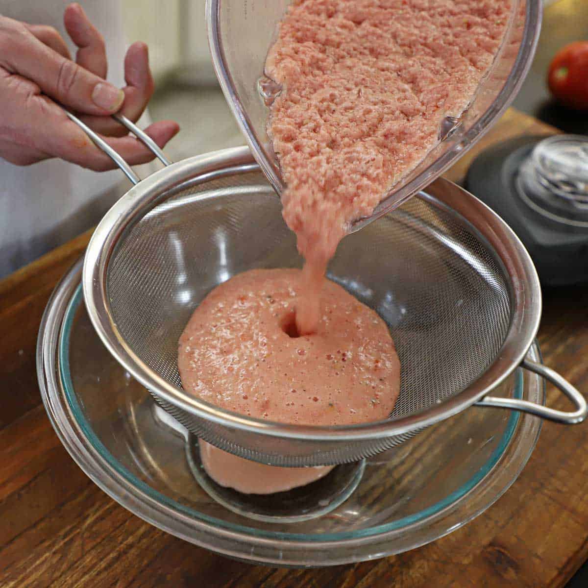 A person passing a puréed tomato and onion mixture through a sieve into a glass bowl.