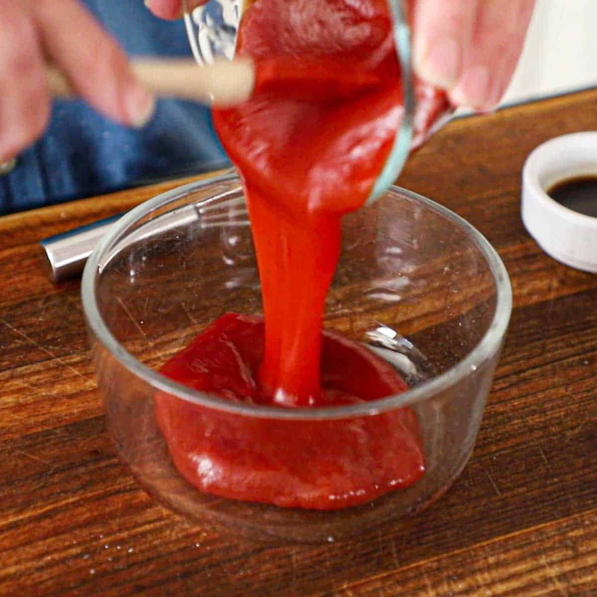 A person pouring ketchup from a small glass bowl into a larger glass bowl on a wooden cutting board.