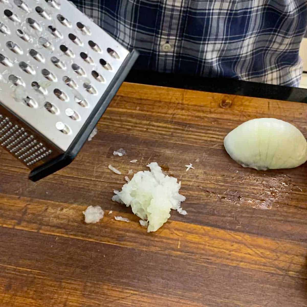 A person lifting a box grated from a wooden cutting board revealing a pile of freshly grated yellow onion.