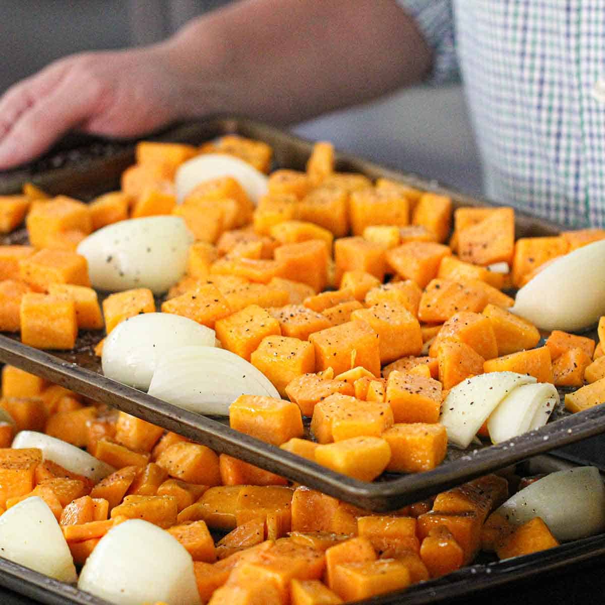 A person holding a baking pan filled with oil and seasoned cubed butternut squash and a quartered onion over a second baking pan filled with the same.