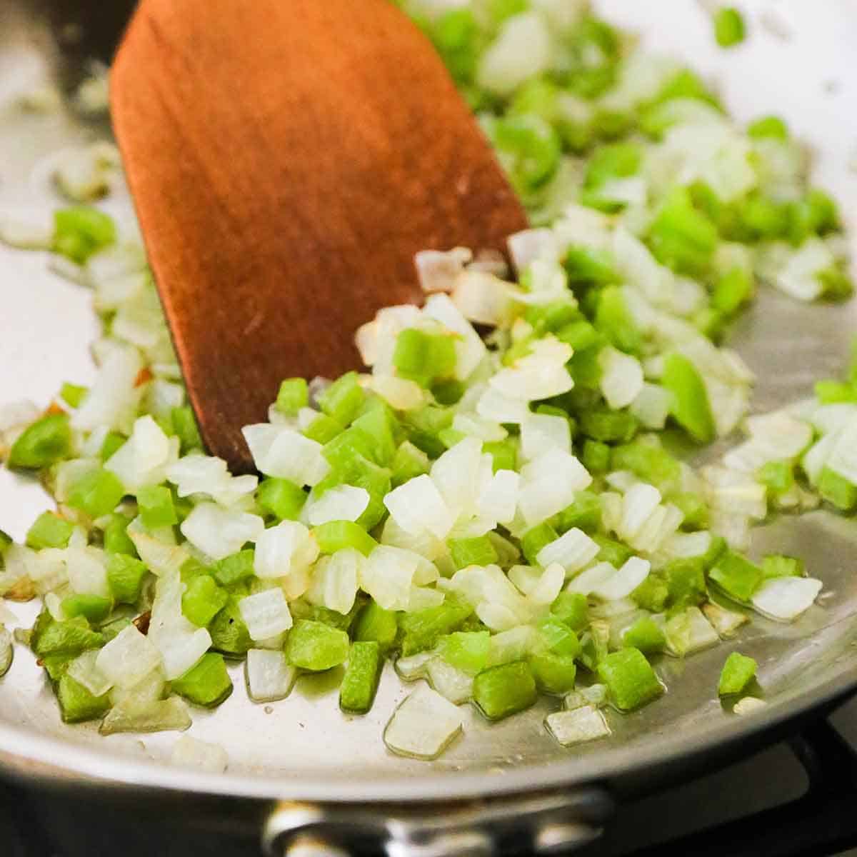 A person using a wooden spatula to stir chopped onion and green bell pepper that is being sautéed in a stainless steel skillet.