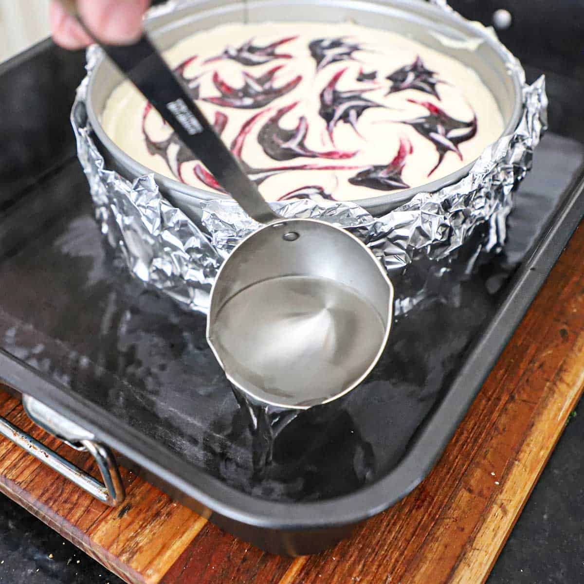 A person using a large ladle to add hot water into a roasting pan that is holding a foil-wrapped springform pan with an uncooked lemon blueberry cheesecake in it.