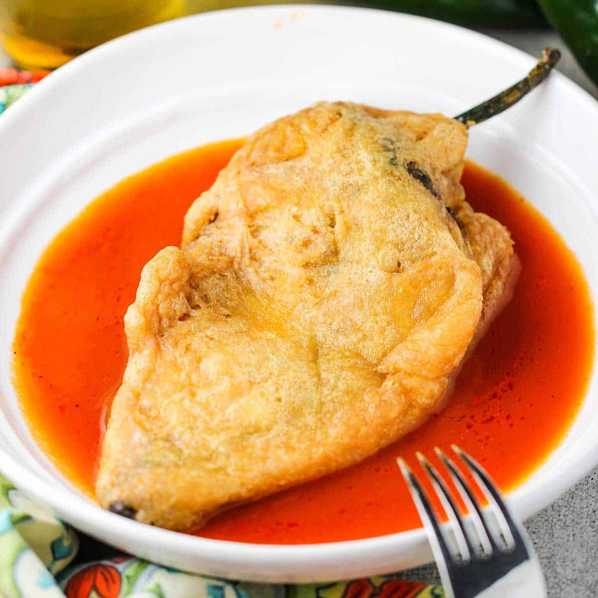 An authentic chile relleno resting in a shallow red sauce in a white bowl.
