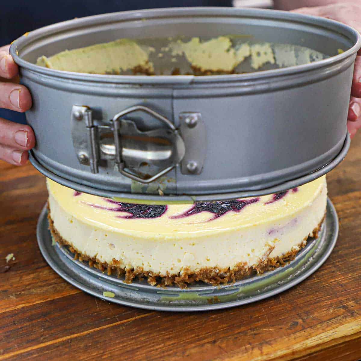 A person lifting a springform pan from a chilled lemon blueberry cheesecake that is resting on a wooden cutting board.