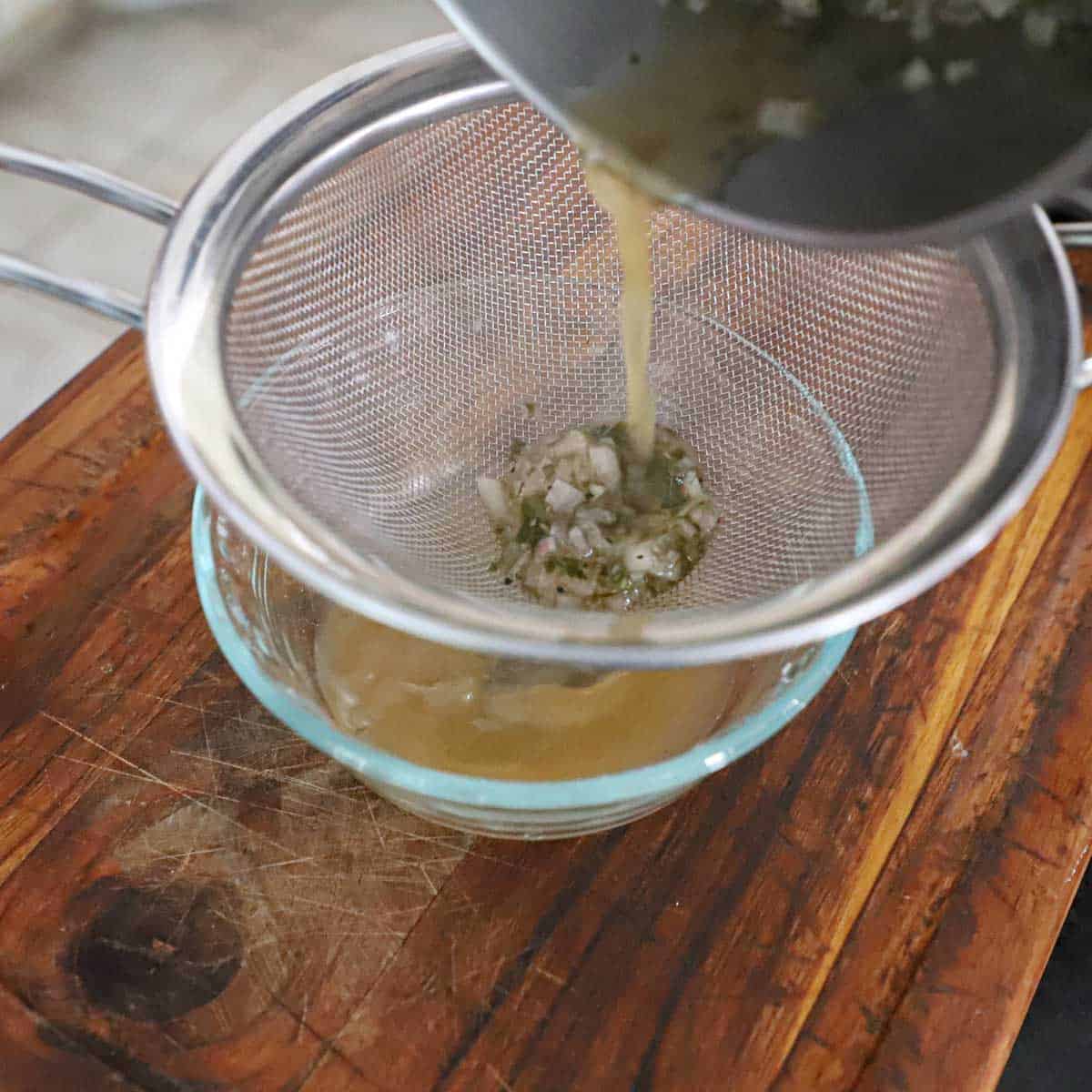 A person straining a tarragon, shallot, and vinegar reduction through a sieve into a small glass bowl.