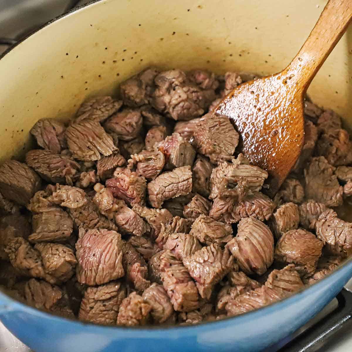 A person using a wooden spoon to stir chunks of chuck roast that are being seared in a large Dutch oven.