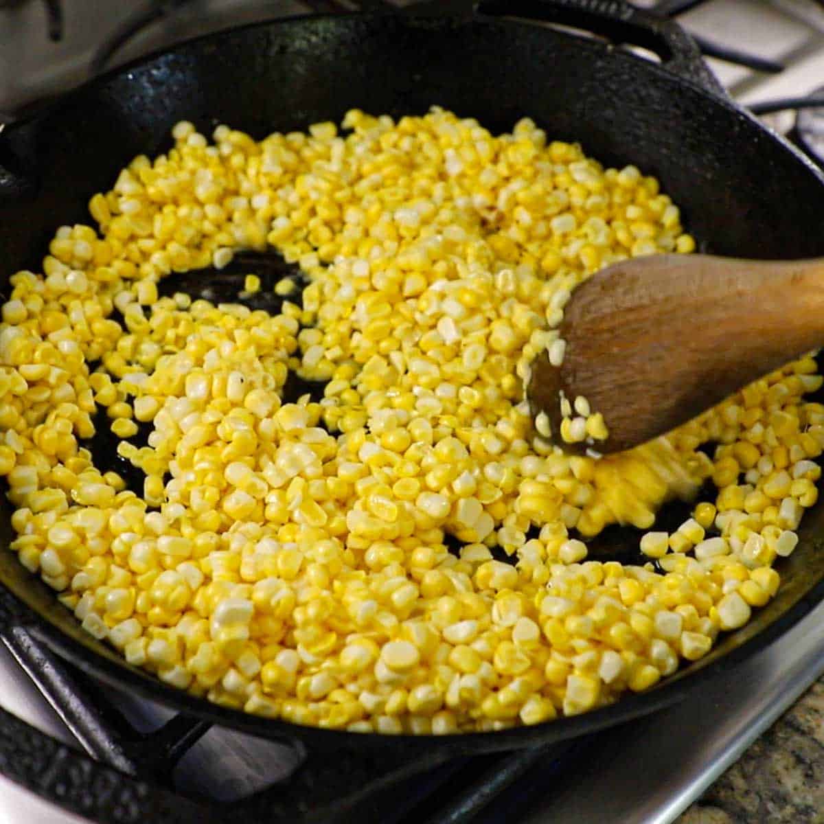 A person using a wooden spoon to cook corn kernels in a large black cast-iron skillet on a gas stove.