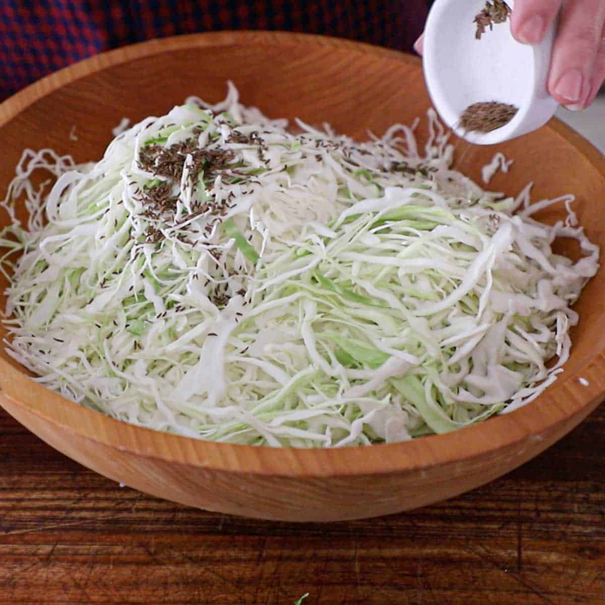 A person transferring caraway seeds from a small bowl into a large wooden bowl filled with shredded green cabbage.