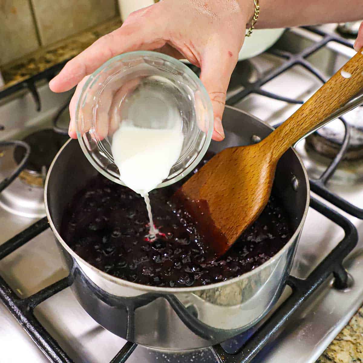 A person pouring a cornstarch slurry from a small glass bowl into a bowl of simmering blueberries that have burst open and created a blueberry sauce.