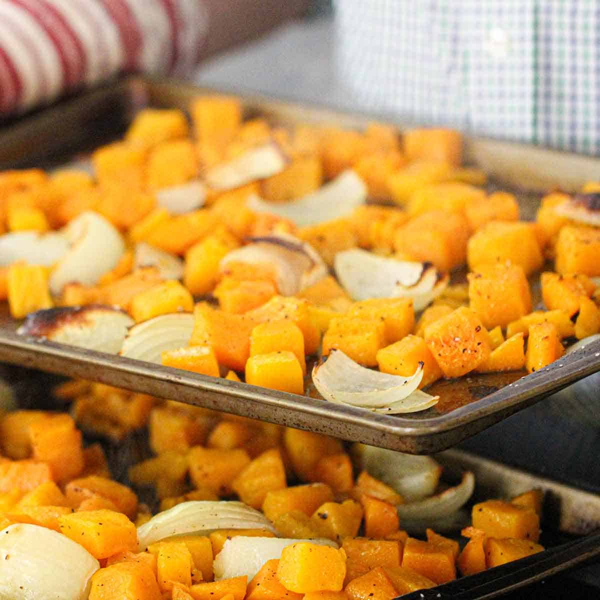 A person holding a baking pan filled with freshly roasted cubed butternut squash and a quartered onion over a second baking pan filled with the same.