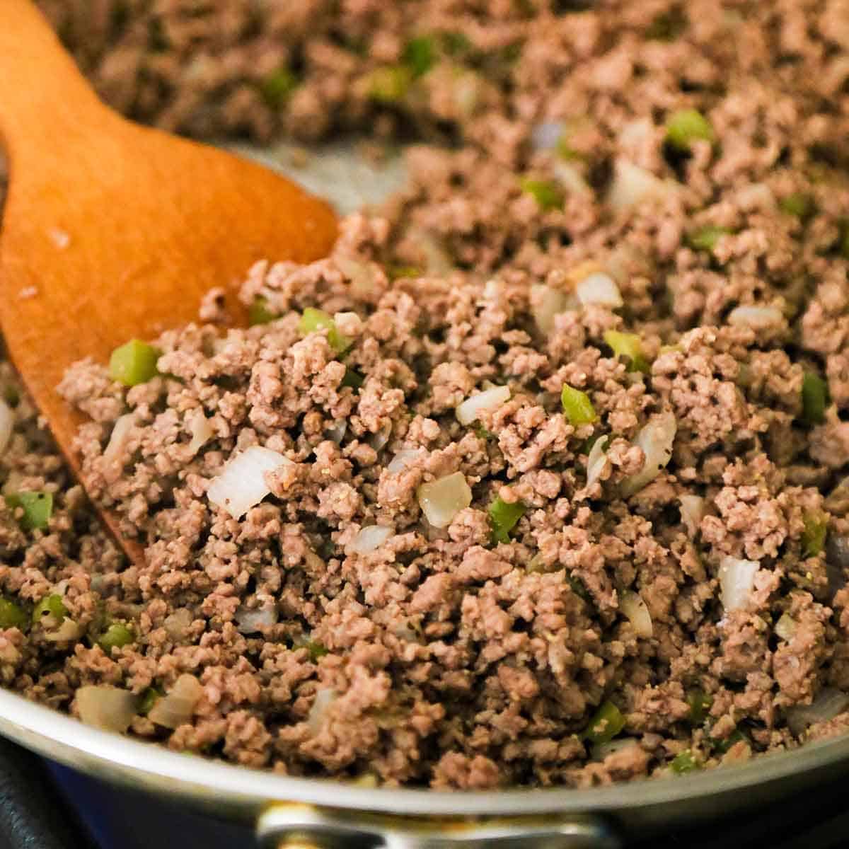 A person using a wooden spatula to stir cooked ground beef with sautéed chopped onion and green bell pepper in a large skillet.