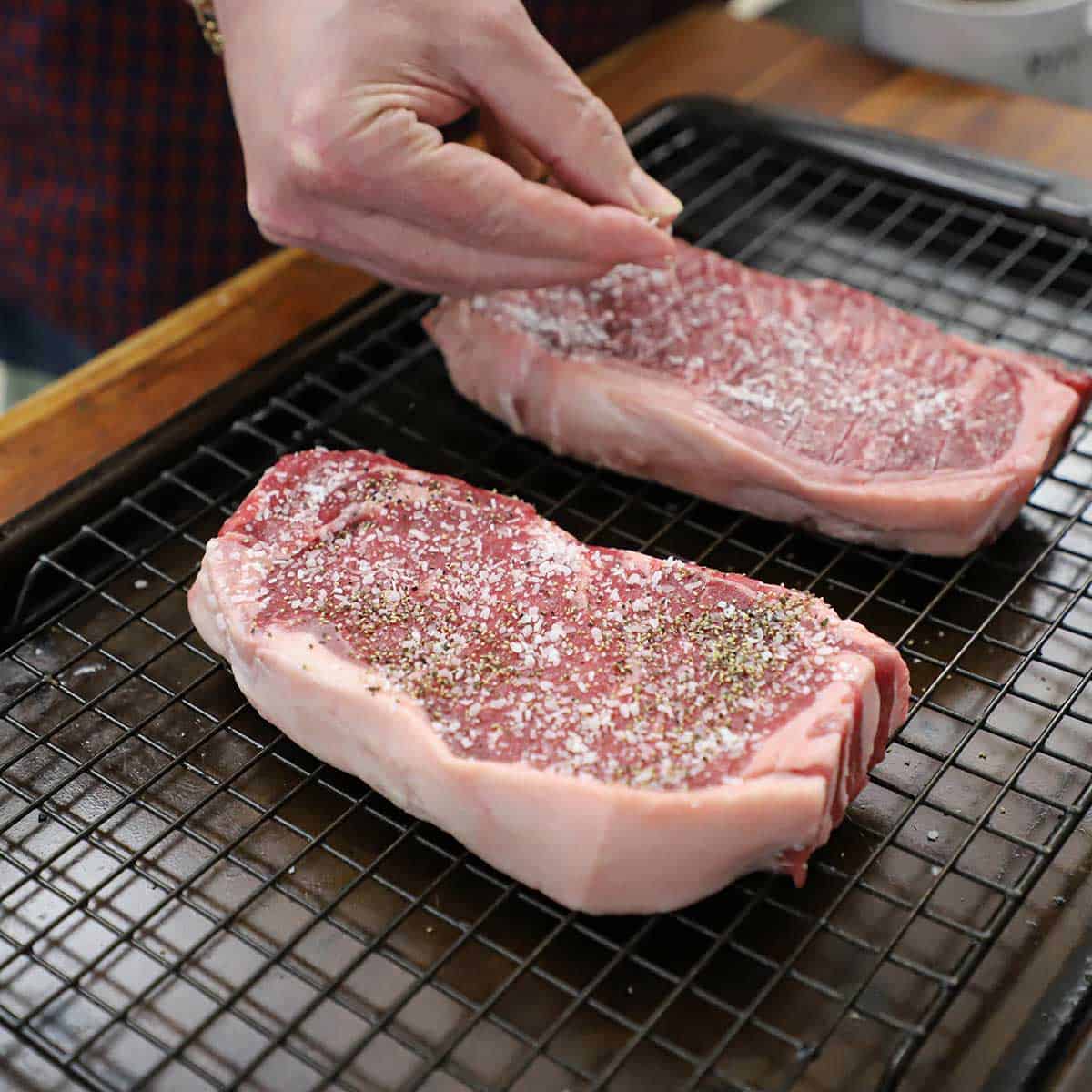 A person using his fingers to sprinkle salt and pepper onto two uncooked strip steaks that are resting on a baking rack on a baking sheet pan.