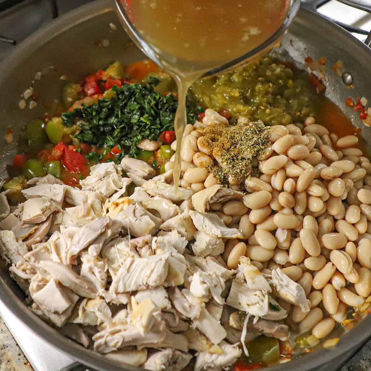 A person pouring chicken stock from a glass measuring cup into a large skillet filled with cannellini beans, cubed rotisserie chicken, sautéed tomatillos, tomatoes, green chiles, and seasonings.